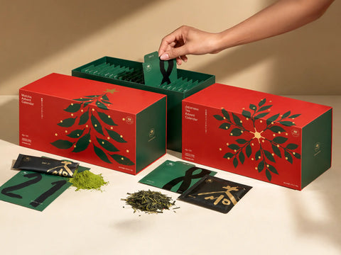 A hand reaches into a green tea gift box while two red and green 'Aurora Borealis' Matcha Tea Collection boxes sit on a beige surface with loose matcha powder and cards in front.