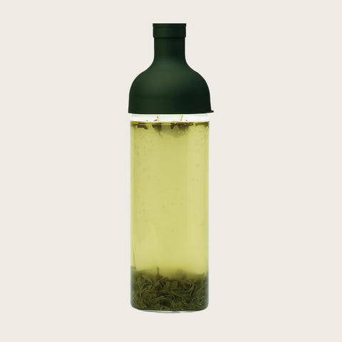 A clear glass bottle with a dark green cap, filled with pale yellow-green tea and showing loose tea leaves at the bottom, set against a solid off-white background.