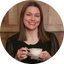 A smiling woman with shoulder-length brown hair holds an antique-style teacup and saucer with gold detailing. She is wearing a black sheer-sleeve top, set against a warm wood-paneled background inside a circular frame.