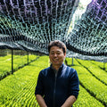 Nakai farmer in a navy blue zip-up sweater stands in a lush green agricultural field under a large black mesh canopy with reflective silver supports.
