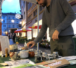 Oliver Staerkle in a gray ribbed cardigan stands at an outdoor market stall, reaching toward small glass jars and packaged items on a table. The scene is set in a real street environment with buildings, market umbrellas, and evening lights in the background.