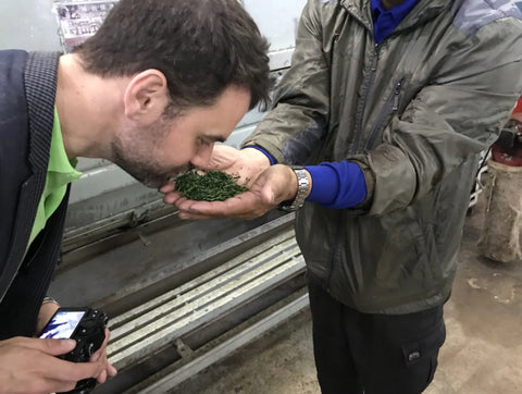 Oliver Staerkle and Sakamoto farmer in an industrial or workshop setting. Oliver Staerkle on the left smells a small pile of green dried herbs or leaves held in the other man's hands.